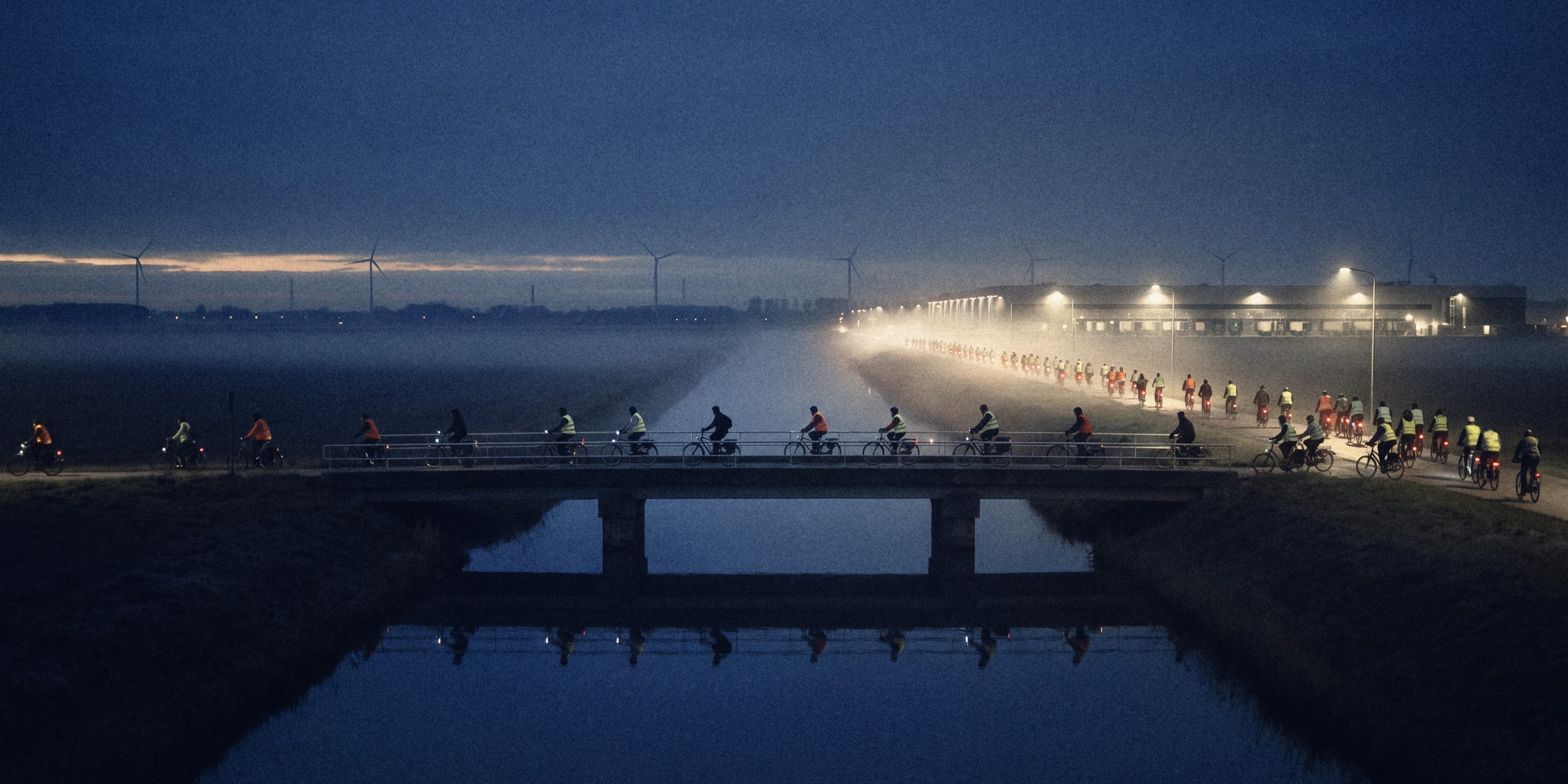 Cyclists in hi-vis cross a canal bridge in the mist at dawn, heading for a lit distribution centre, wind turbines on the horizon