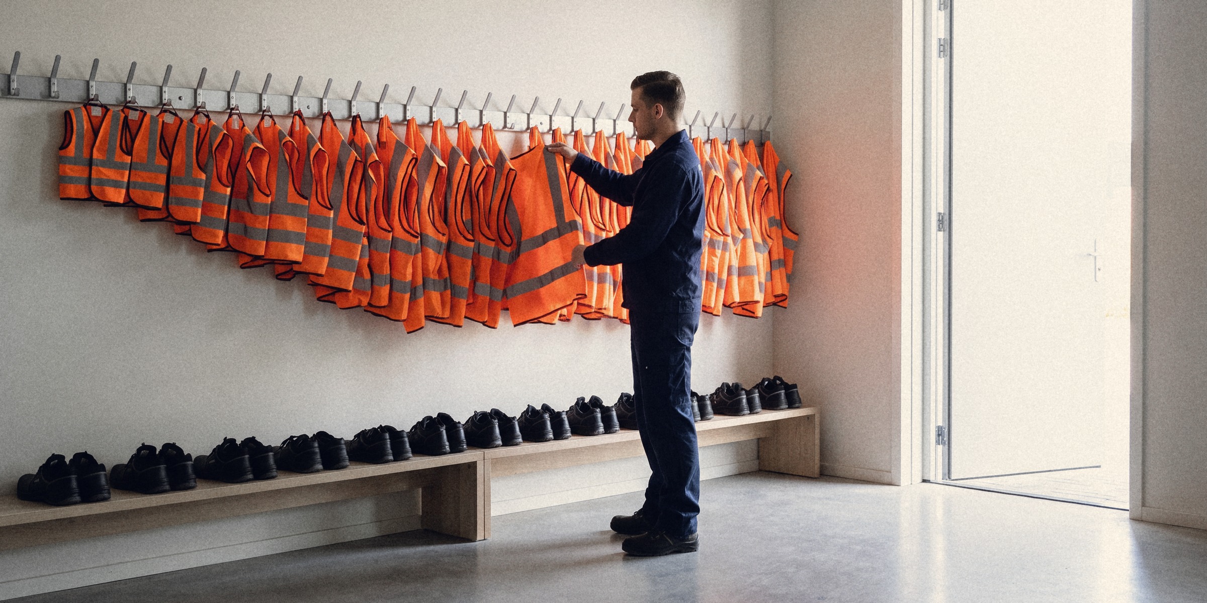 Orange hi-vis vests in every size hang on a row of hooks above safety shoes in every size; a new starter takes one from the middle