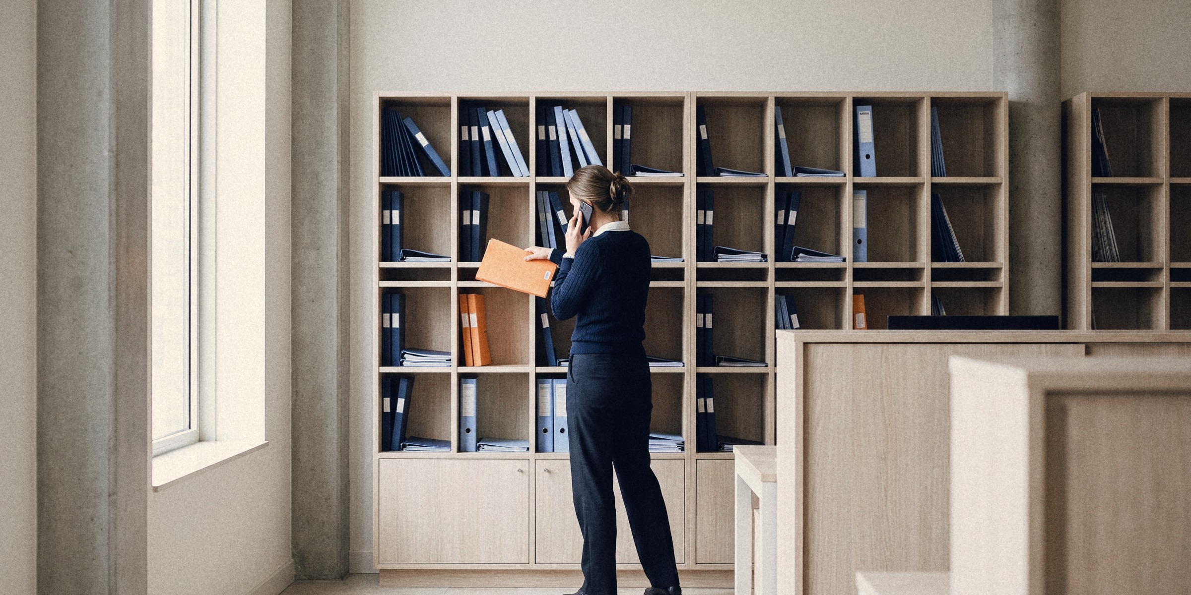 Een planner van een uitzendbureau schuift al bellend een oranje map in een wand van eiken vakken met marineblauwe en lichtblauwe klantmappen