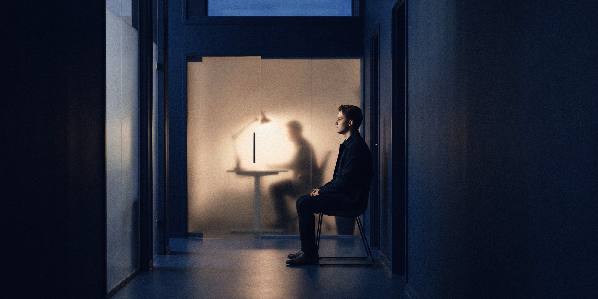 A young applicant waits in a dark corridor facing a frosted glass door, a person working at a lamp-lit desk behind it