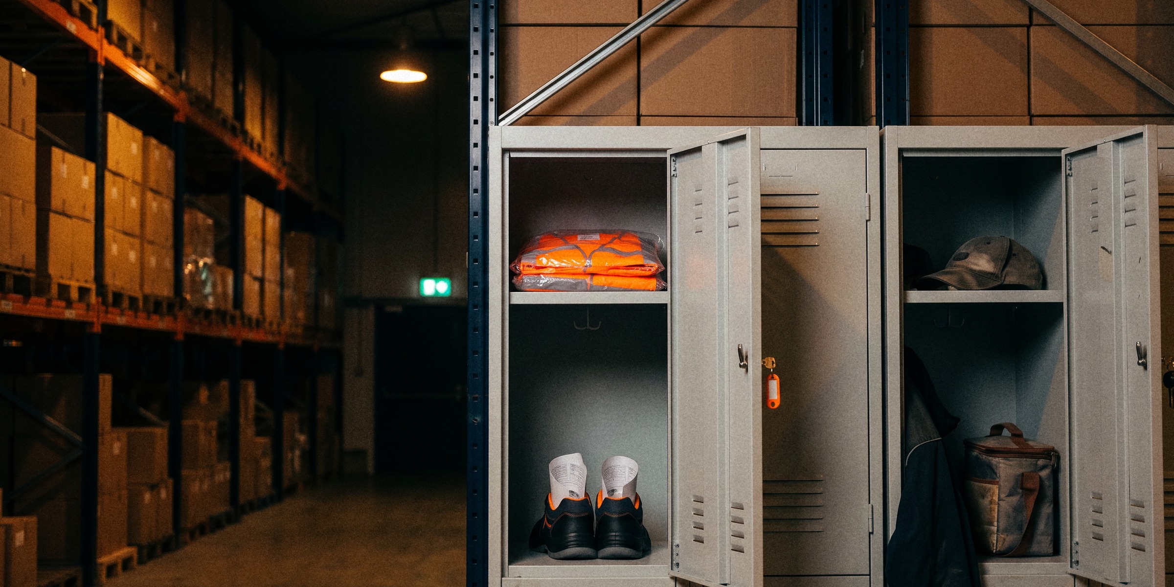 Een open kluisje in een donker magazijn met een nieuw hesje nog in het plastic en nieuwe werkschoenen, de sleutel met een oranje label nog in het slot