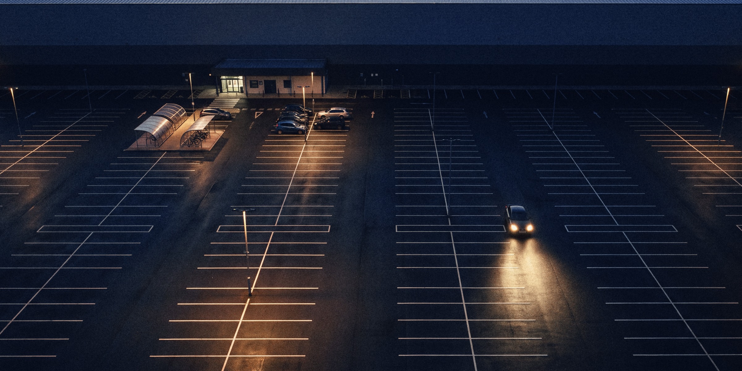 A drone view at dawn of a distribution centre's staff car park, most bays empty, one car arriving with its headlights on