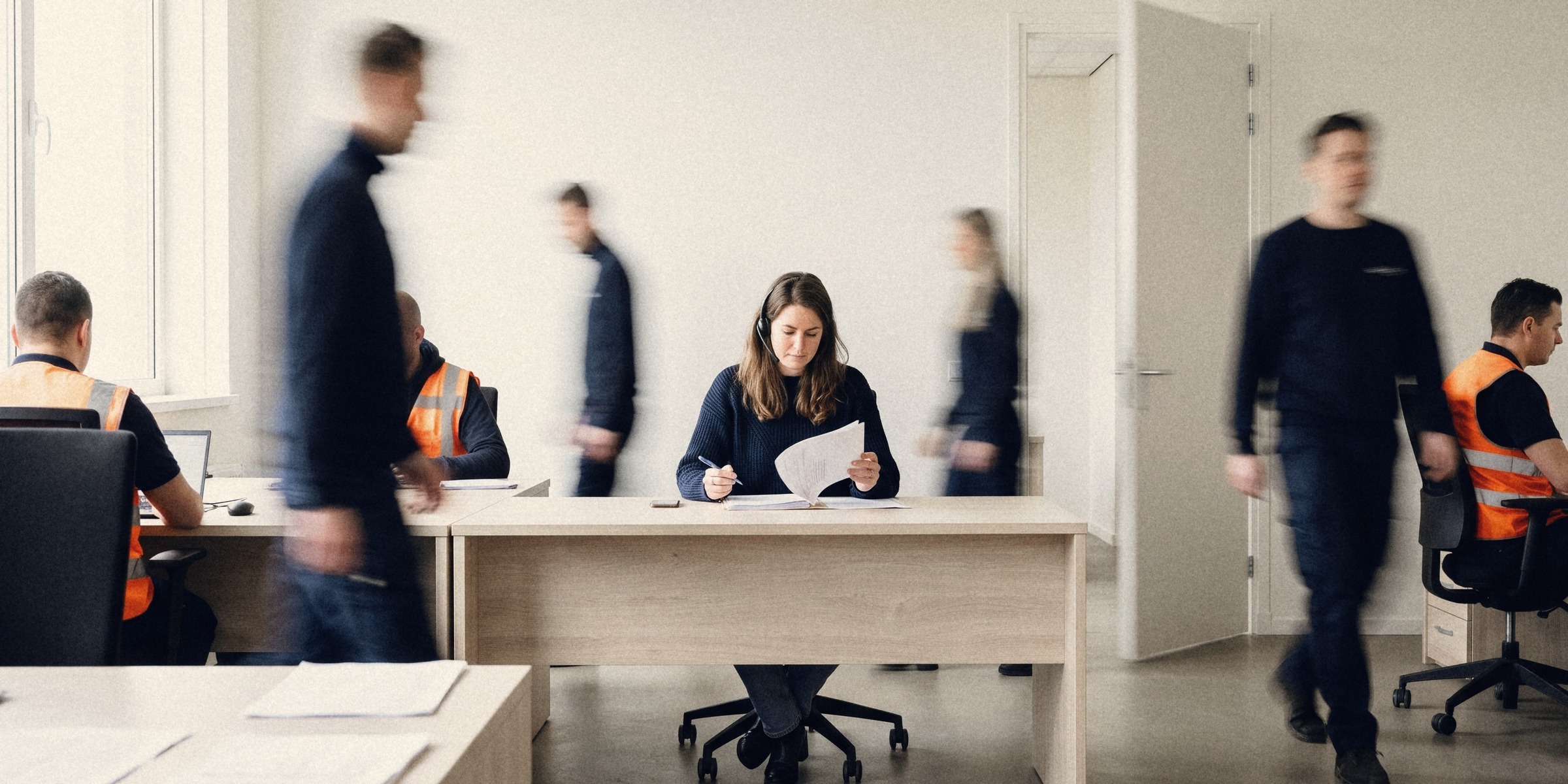 In een druk kantoor van een uitzendbureau, als lange sluitertijd vastgelegd, lopen collega’s wazig voorbij terwijl één intercedent stil een kandidaatdossier leest