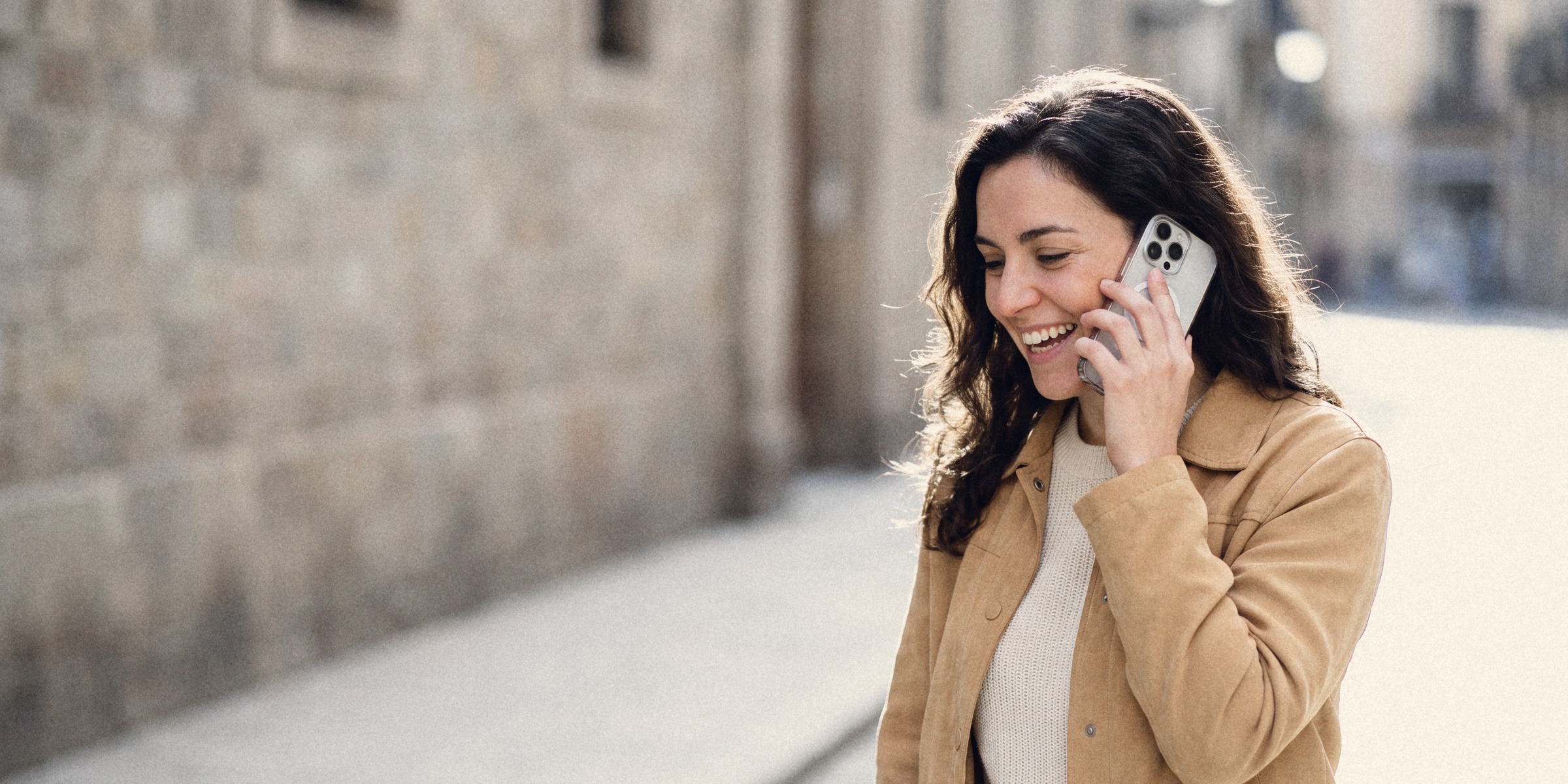 A young woman in a tan suede jacket laughing on a phone call outdoors