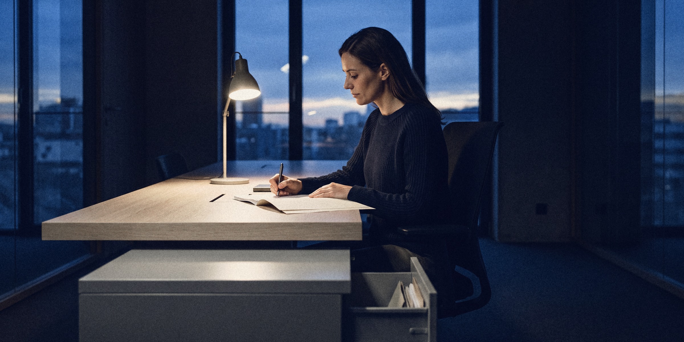 A recruiter writing a note into a candidate file under a desk lamp at blue hour