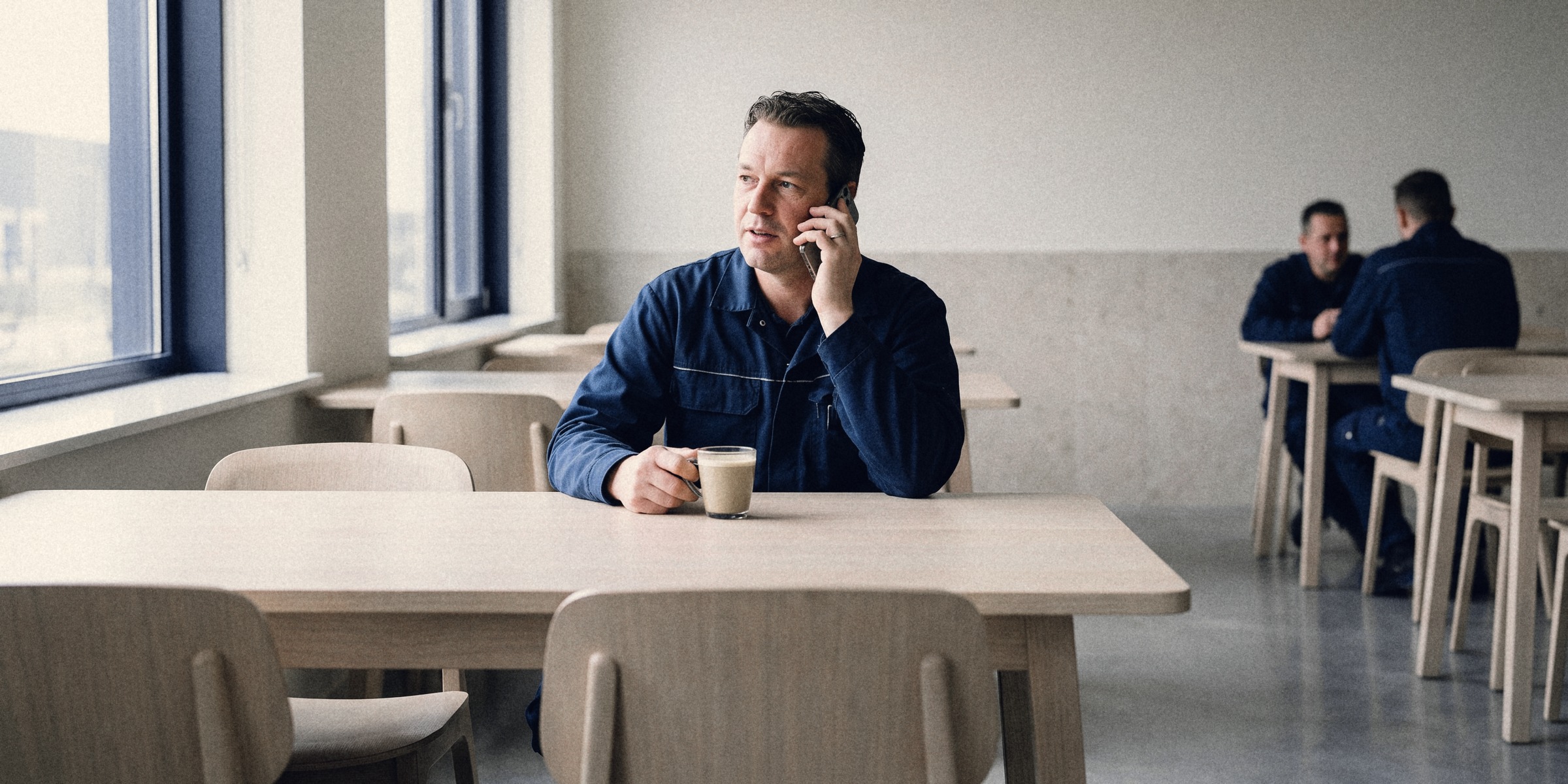 A man in a navy work jacket on a phone call at a canteen table by the window