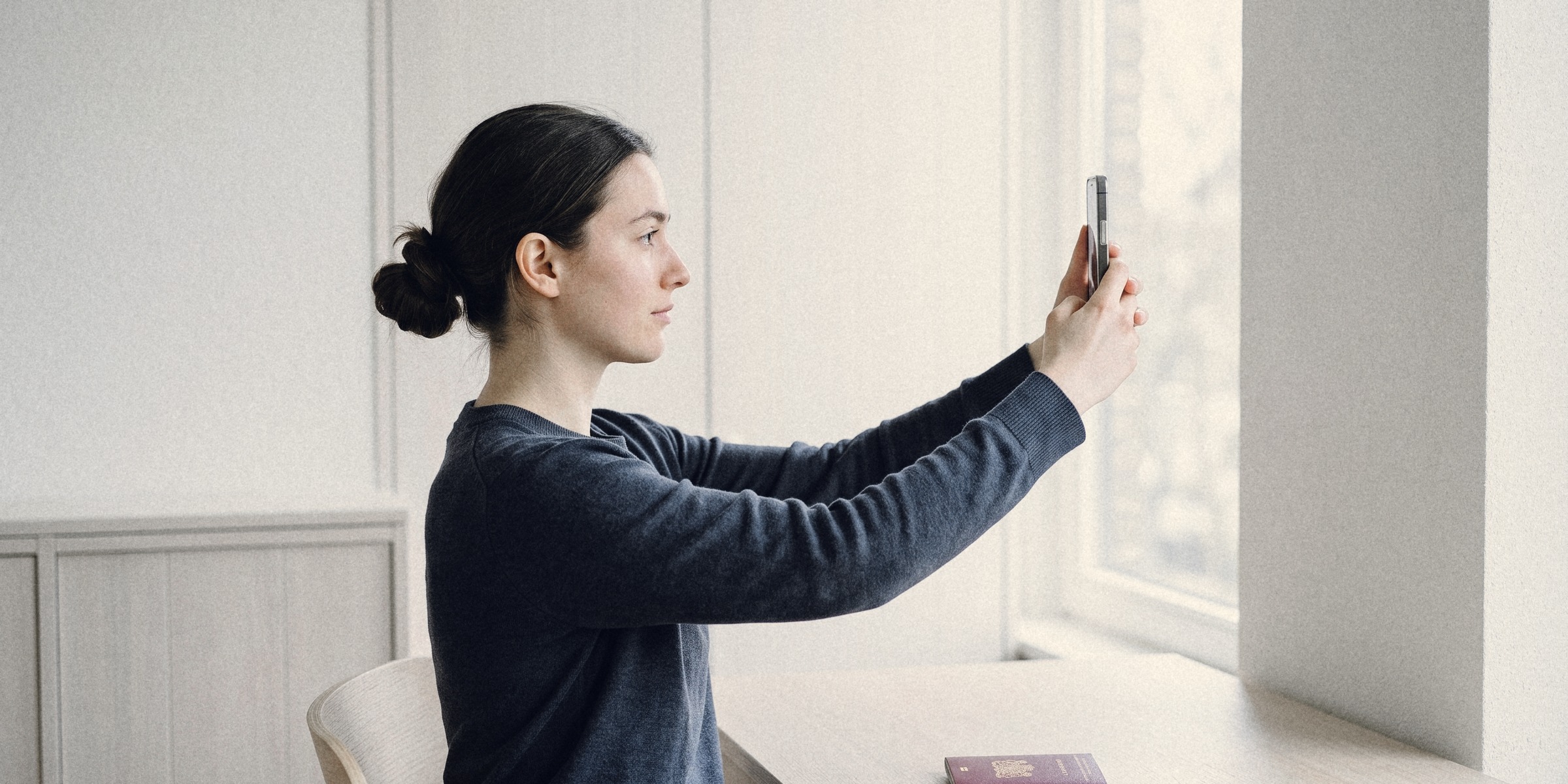 A woman at a desk taking a selfie at arm's length, a closed passport in front of her