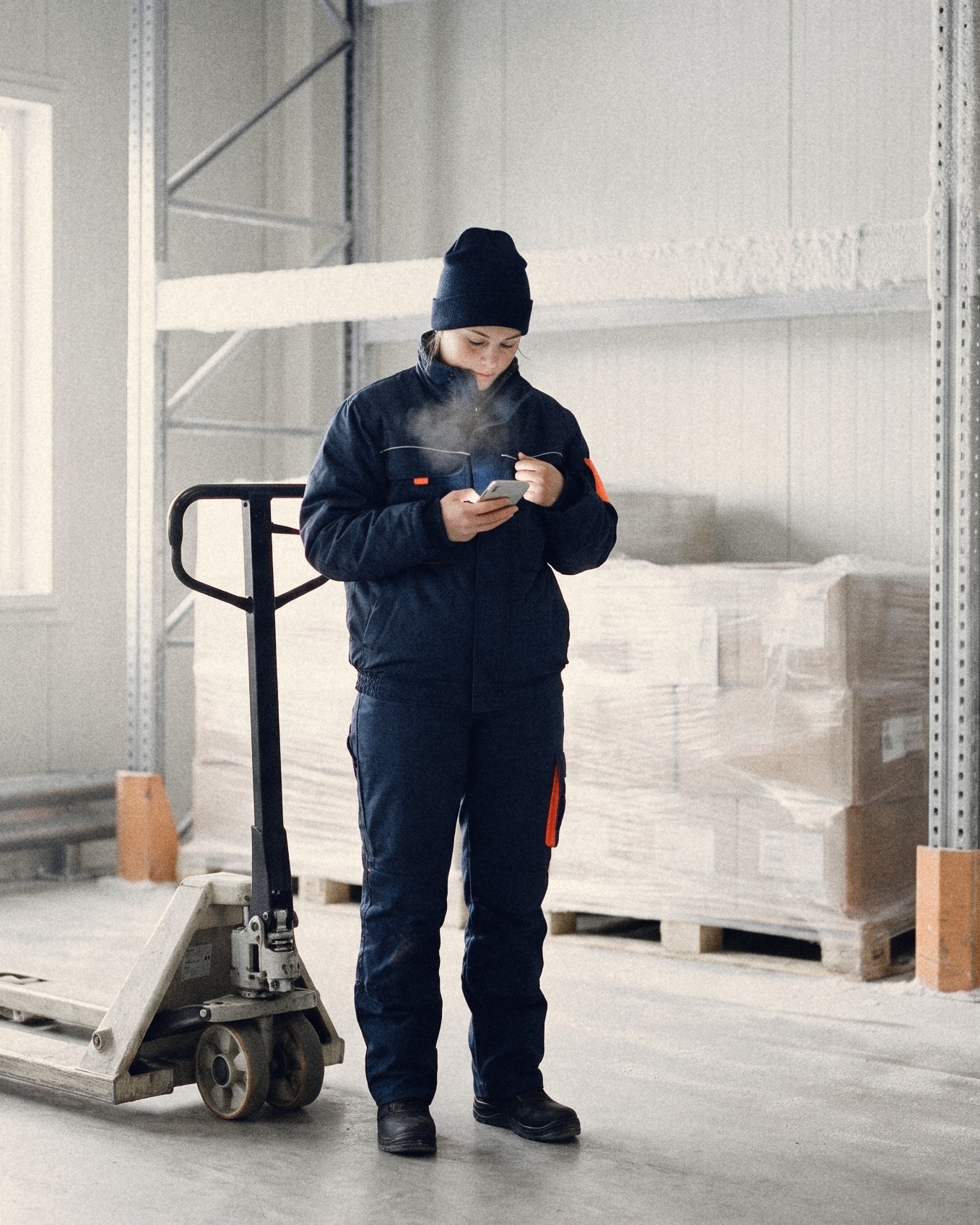 A worker in a navy freezer suit checks a text on her phone in a frosty cold store, her breath visible, a pallet truck beside her
