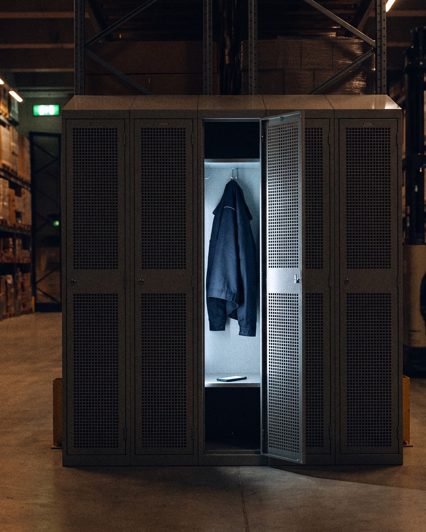 An open locker on the warehouse floor holds a jacket and a lit phone while its owner drives a reach truck in the background