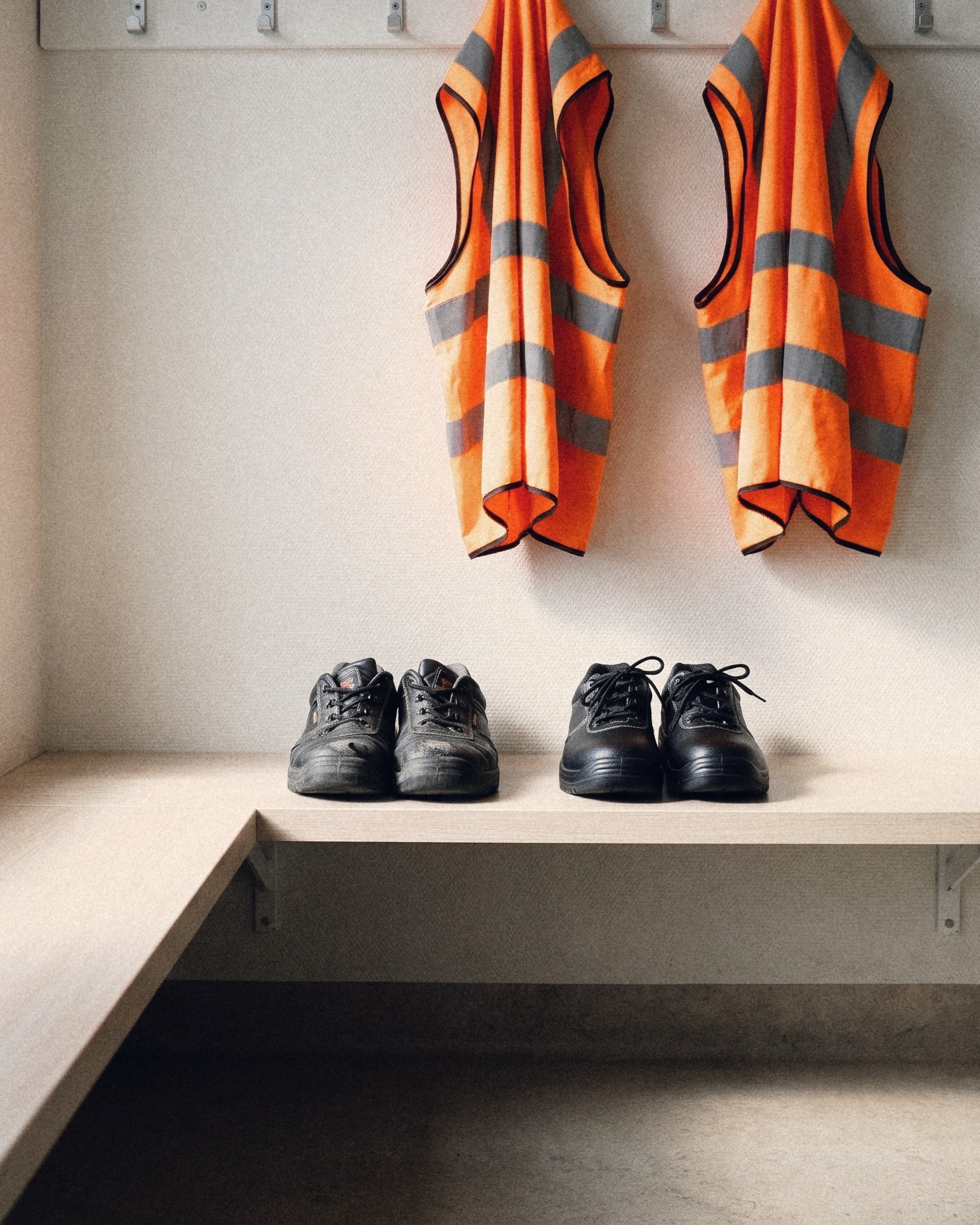 A worn pair and a brand-new pair of safety shoes side by side on a locker-room bench, two hi-vis vests on the hooks above