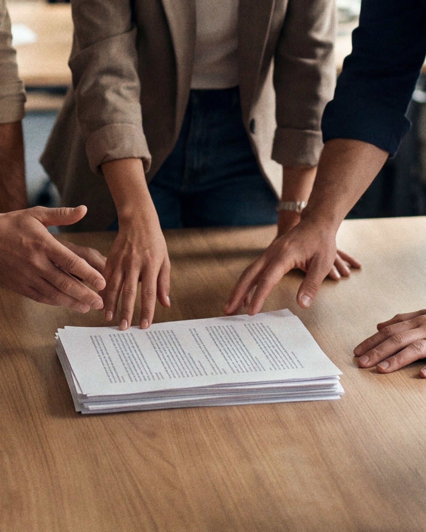 Hands of a team reaching toward one stack of printed papers on an oak table