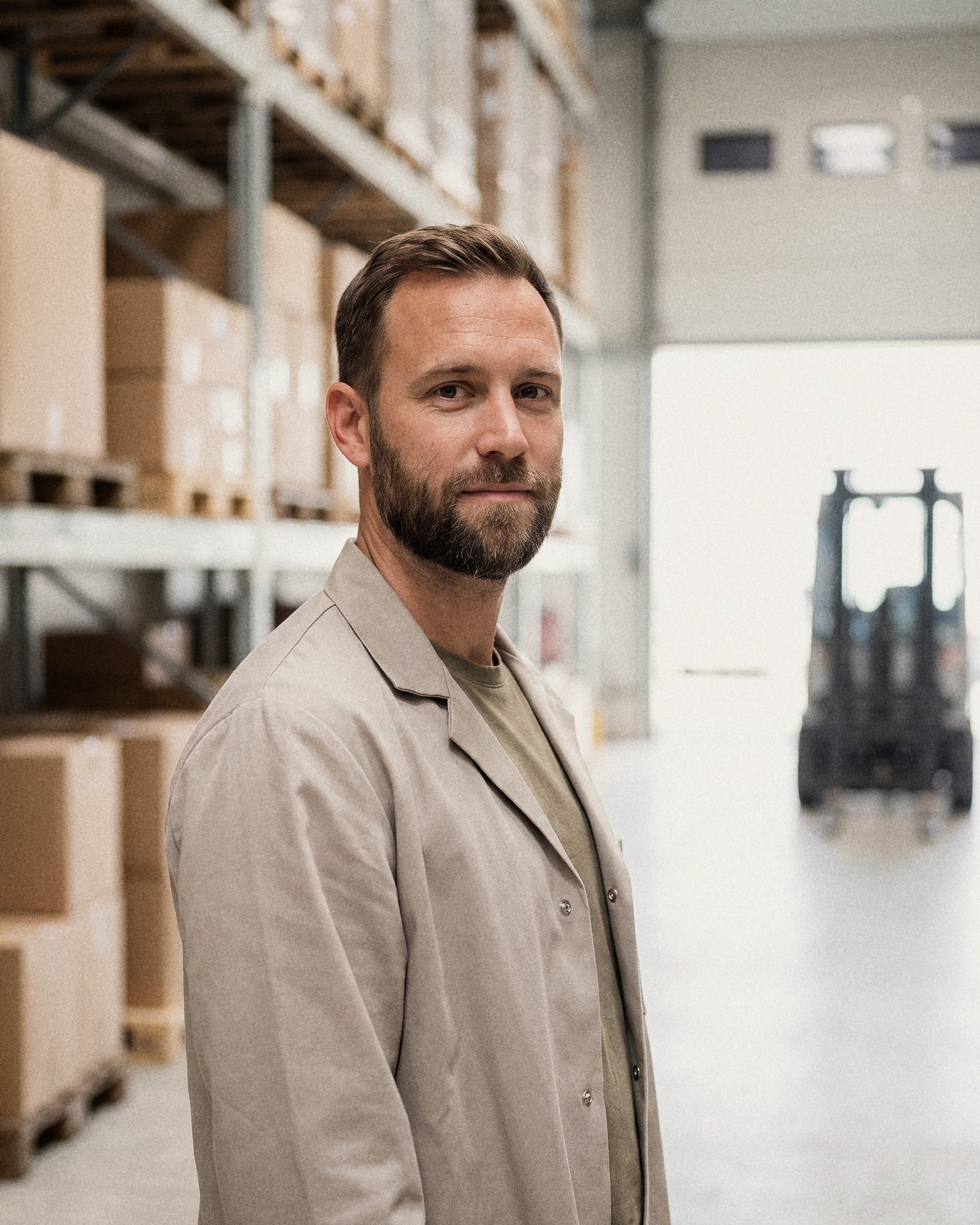 A bearded warehouse worker in a beige work coat in an aisle