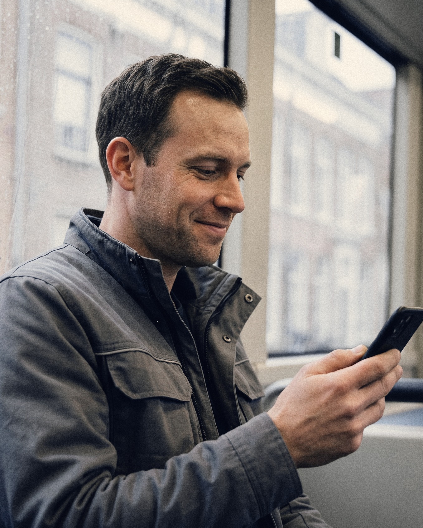 A man on a bus reading a message on his phone with a small smile, rain on the window