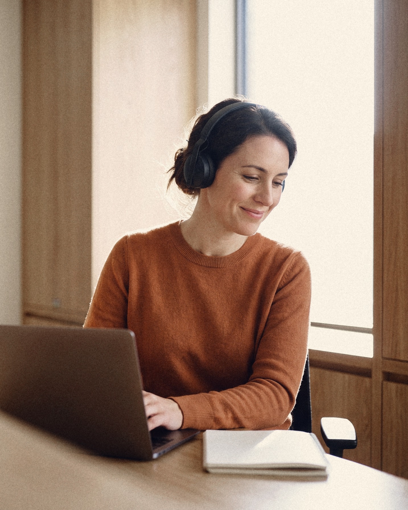 A woman with headphones typing on a laptop beside a notebook